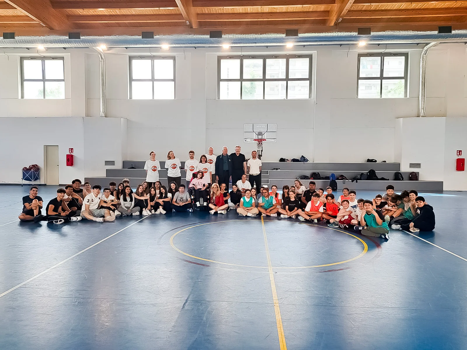 Grande gruppo di atleti seduti in cerchio sul parquet, generazioni unite